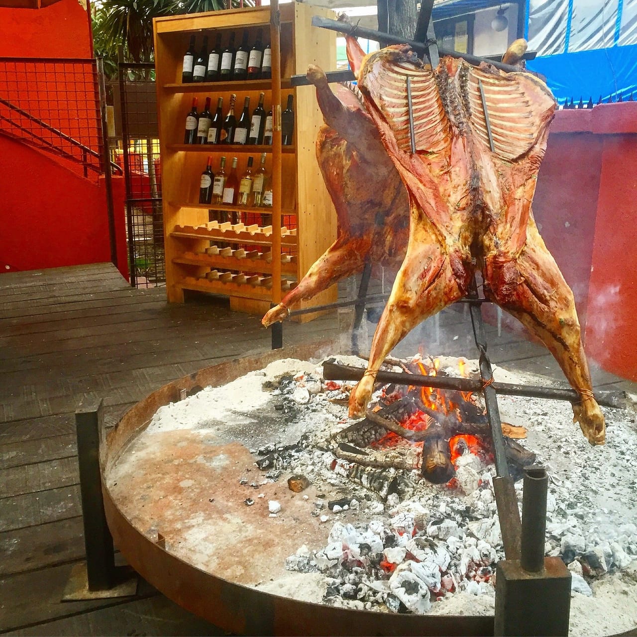Piezas de carne abiertas en cruz asándose junto a un fuego de leña, con botellero de vinos al fondo