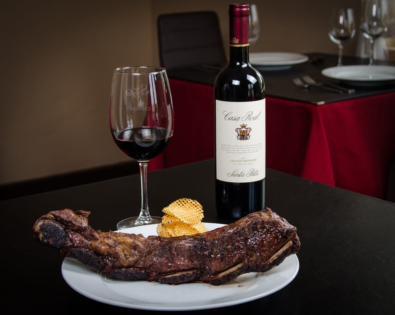 Corte de carne a la brasa sobre plato blanco, junto a una copa de vino tinto