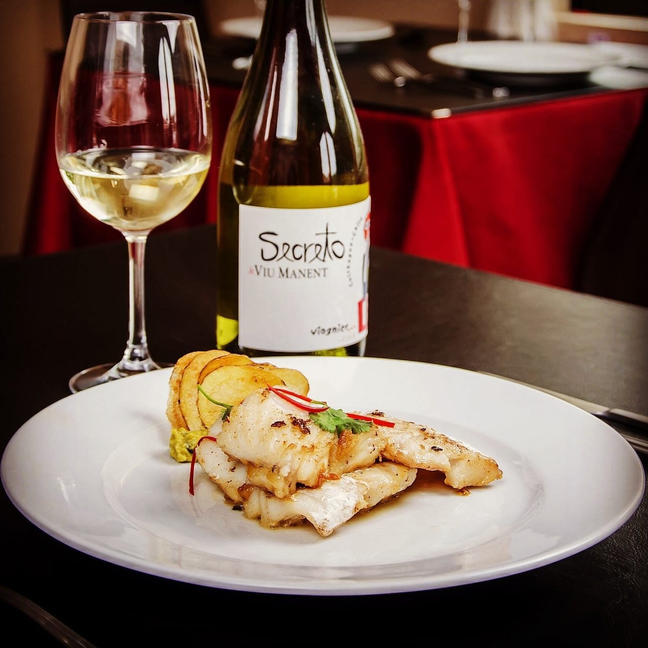 Lomo de pescado blanco con láminas de papa, junto a una copa de vino blanco
